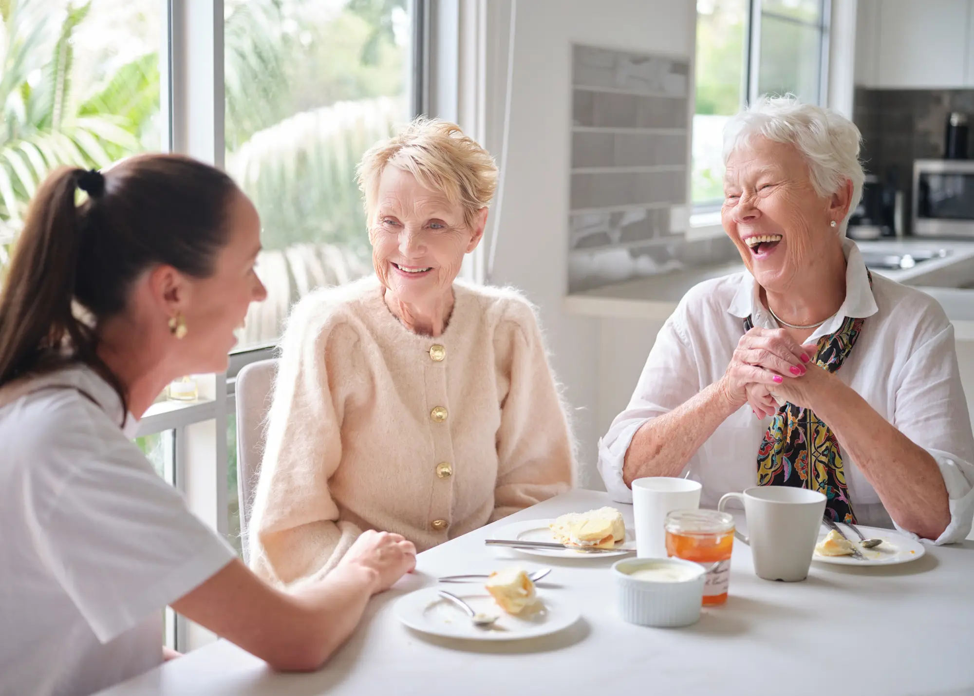 A home carer sharing afternoon tea and a laugh with two older women at a sunny kitchen table.